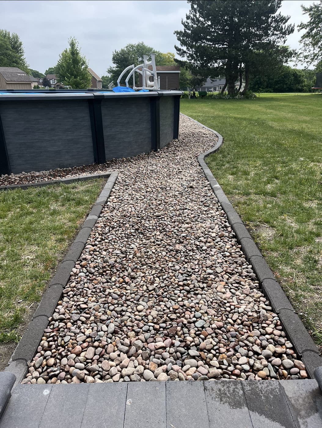 Gravel pathway beside a swimming pool in a landscaped yard with trees.