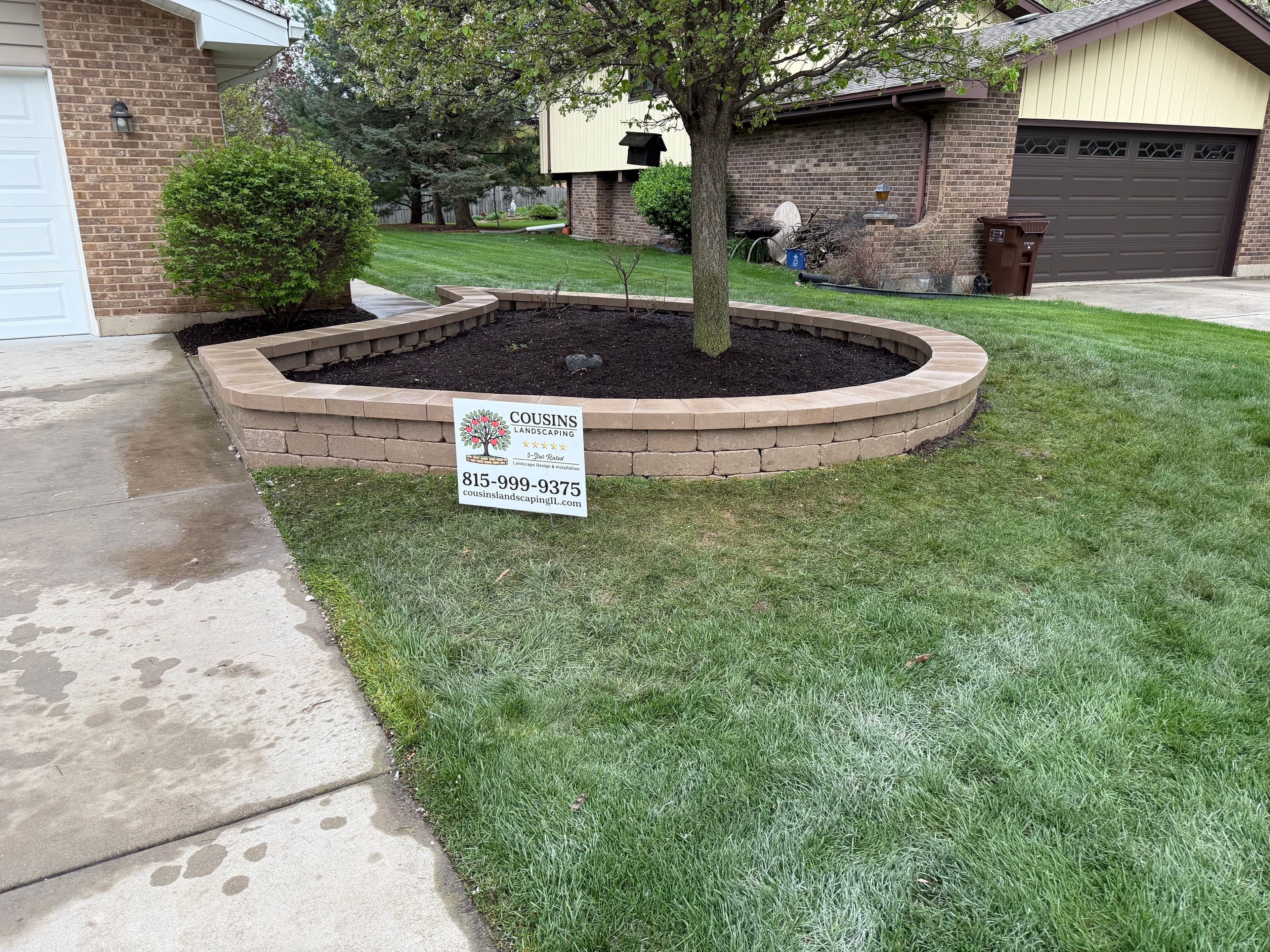 Landscaped yard with a tree and stone border, featuring a sign for Cousins Landscaping.