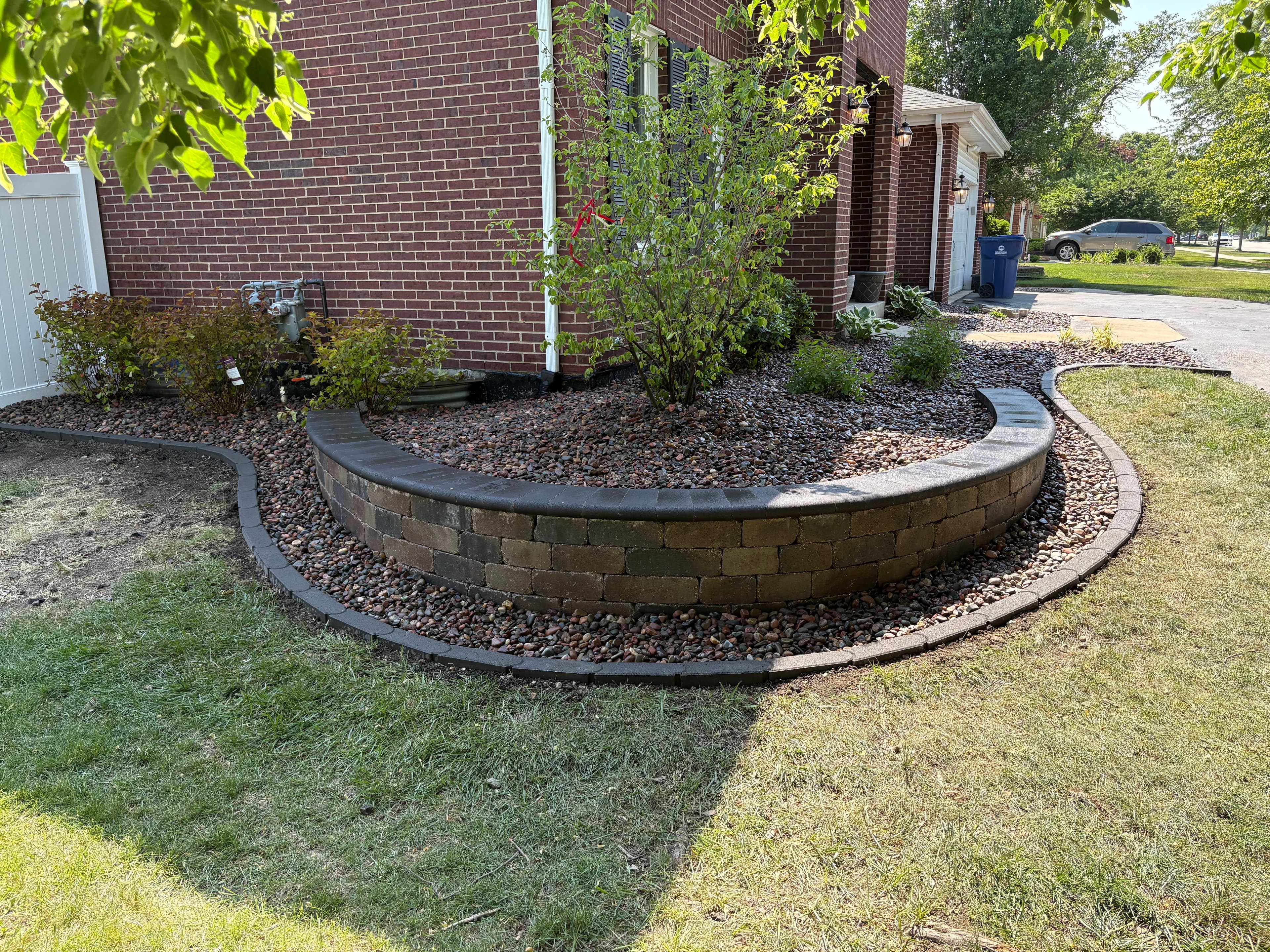 Curved stone retaining wall in a landscaped yard with gravel and green plants.