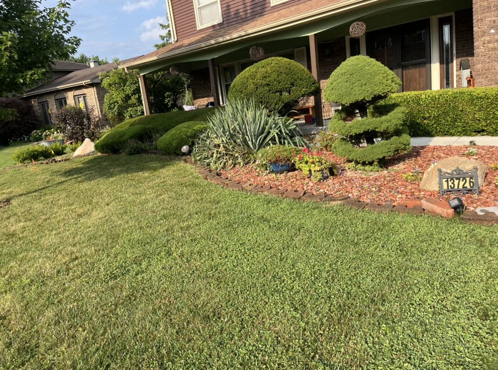 Lush front yard landscaping with green shrubs, flower beds, and a house number sign.