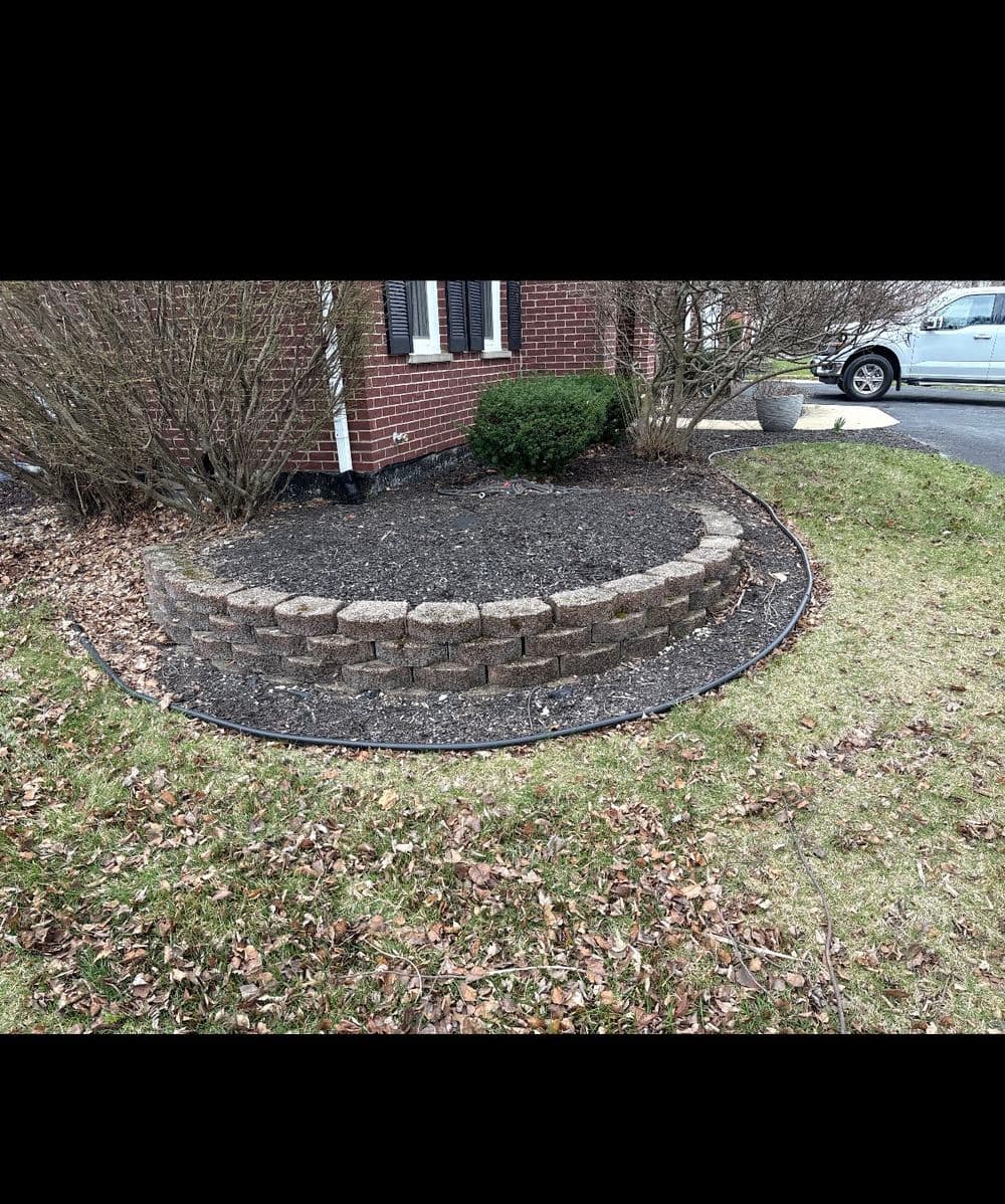Curved stone retaining wall in garden with mulch and bare patches, surrounded by grass.